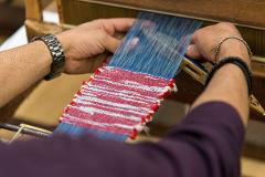A person's hands are shown weaving a red and white striped fabric on a loom with blue threads.