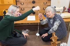 Two women are sitting on the floor, one showing the other how to spin thread on a traditional charkha.