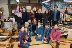 A group of people, some kneeling and some standing, pose for a photo in a workshop with traditional weaving equipment and products.