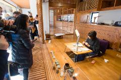 People in a room with wooden walls and floors observe a woman working at a table with a lamp and various items.