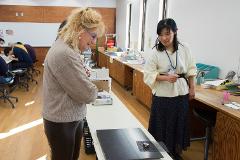 Two women, one with blonde curly hair and the other with black hair, stand by a table in a brightly lit room with wooden paneling.
