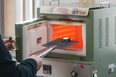 A person's hand holds a small metal object on a grate, carefully placing it into a glowing orange furnace.