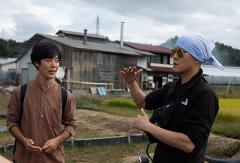 Two men are talking in front of a building with a cloudy sky in the background, with one man wearing a bandana and sunglasses.