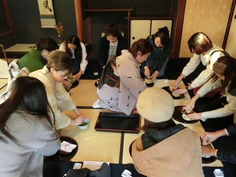 A group of people are gathered in a traditional Japanese room, seated on tatami mats, participating in a tea ceremony. They are dressed in both traditional and modern attire, preparing and enjoying matcha tea.