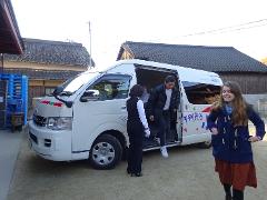 A white van is parked outside a building while a man steps out of the van and a woman stands nearby, with another woman standing off to the side.