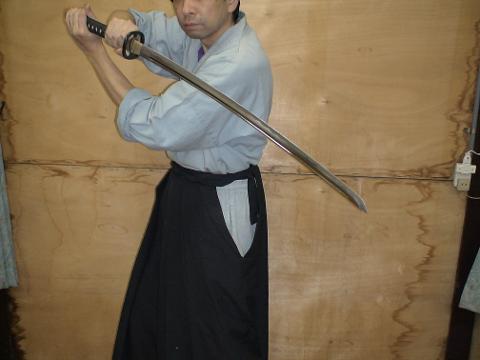 A person in a light gray shirt and black pants is holding a katana sword.