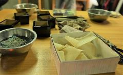 Use Premium Shark Skin and Traditional Lacquer to Craft a Masu Cup near Inuyama Castle A set of tools and materials, including bowls, black containers, scissors, and a box of textured material, are laid out on a wooden table, likely for crafting a masu cup.