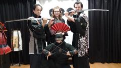 Um grupo de cinco pessoas vestidas com vários trajes tradicionais japoneses, incluindo um samurai e um ninja, posam com espadas e um leque em frente a uma cortina escura.