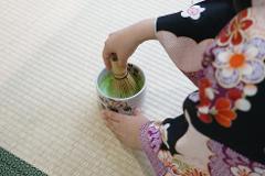 A person in a traditional Japanese kimono whisks matcha tea in a decorative bowl while seated on a tatami mat.