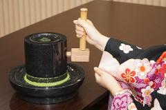 A woman in a kimono grinds tea leaves with a wooden pestle in a stone mortar.