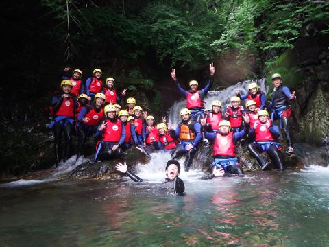 A group of at least 20 people in wetsuits, helmets and red life jackets pose for a photo in a river with a small waterfall behind them. One person in the water screams with their arms outstretched.
