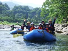 Eine Gruppe von Menschen genießt eine Wildwasser-Raftingtour auf einem Fluss. Sie tragen Helme und Schwimmwesten und heben aufgeregt die Hände.