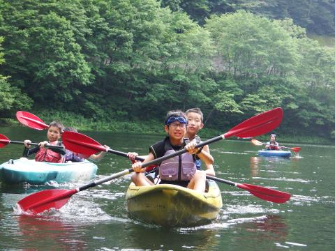 Several children are kayaking on a calm lake surrounded by lush green trees.