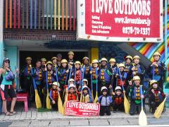 A group of people, including children, wearing life vests and helmets, holding paddles, pose in front of a building with an "I Love Outdoors" sign.