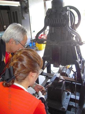 Two people are watching a metal shaping machine in operation, possibly blacksmithing or metalworking.