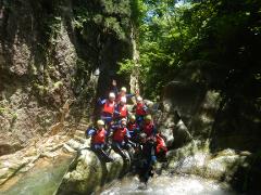 A group of ten people wearing wetsuits, life vests, and helmets pose for a photo on rocks in a river, surrounded by lush greenery and rock walls.