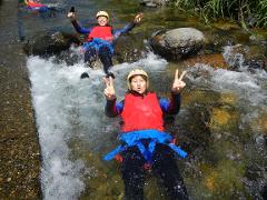 Two people in wetsuits and helmets float in a rushing river, making peace signs, while another person is visible in the background.