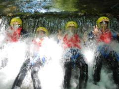 Four people in helmets and wetsuits pose under a waterfall, partially obscured by the cascading water, with two of them making peace signs.