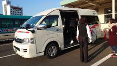 A white van with an open door is parked on a sunny day, as a person steps inside and two women stand nearby.