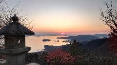 A serene sunset view from a hillside featuring a stone lantern, mountains, islands, and a calm body of water reflecting the orange and pink sky.