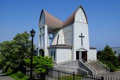A full shot of a white church with a unique brown roof design on a sunny day, featuring stairs leading to the entrance and surrounding greenery.