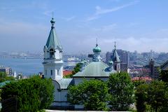 A scenic view of the Hakodate Orthodox Church with its distinctive architecture, set against a backdrop of a cityscape, sea, and hazy blue sky.