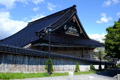 A Japanese-style building with a dark tiled roof stands behind a textured wall topped with matching tiles, lit by a black street lamp under a bright blue sky with scattered clouds.