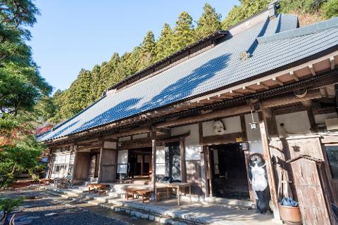 A traditional Japanese building with a tiled roof, wooden beams, and sliding doors, nestled amongst lush green trees under a clear blue sky.