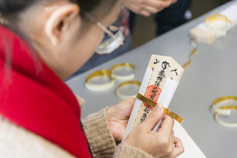 A person with glasses and a red scarf is carefully handling a wooden plaque with Japanese writing, secured with a golden ribbon, while sitting at a table with several white and gold rings.