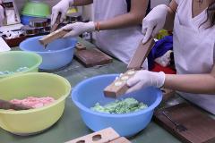 Eldiven ve beyaz önlük giymiş iki kişi, plastik kaplardaki renkli hamurları tahta kalıplarla şekillendiriyorlar.