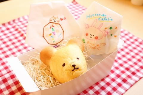 A box containing a bear-shaped pastry sitting on straw, flanked by two paper bags with cartoon drawings, all placed on a red and white checkered cloth.