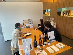 A group of people are gathered around a wooden counter, with bottles of sake and pamphlets displayed on the surface.
