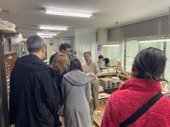People gather in a pottery workshop, with shelves of ceramics and windows showing a bright sky.