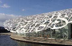 A building with a unique, white Arabic-style canopy sits by the water under a blue sky.