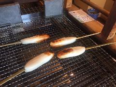 Four skewers of mochi, a Japanese rice cake, are being grilled over a metal grate.