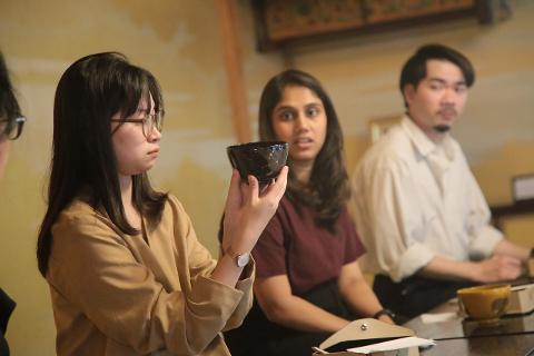 Discover the Art of Yamanaka Lacquerware & Experience the Ultimate Hospitality of the Tea Ceremony Three individuals are seated around a table, with the woman in the foreground examining a dark lacquered bowl, likely during a tea ceremony.