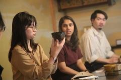 Discover the Art of Yamanaka Lacquerware & Experience the Ultimate Hospitality of the Tea Ceremony Three individuals are seated around a table, with the woman in the foreground examining a dark lacquered bowl, likely during a tea ceremony.