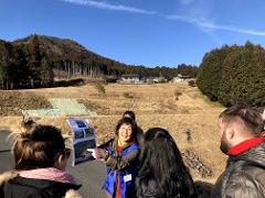 A group of people is looking at photos in front of terraced fields and a forested mountain under a clear blue sky.