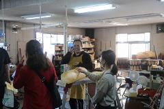 A smiling man in an apron talks on his phone while a woman examines a large, curved wooden piece in what appears to be a workshop filled with drums and supplies.