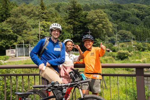 Three men on bicycles ride through a forest on a dirt path.