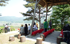 People are gathered under a wooden pavilion overlooking a valley, some observing a distant scene, possibly at a pottery event.