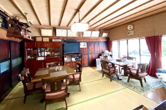 A traditional Japanese dining room features tatami flooring, wooden furniture, and large windows with red curtains, offering a glimpse of an outdoor scene.