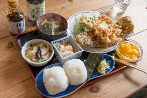 A Japanese meal set is presented on a black tray, including rice balls, fish, shredded cabbage, soup, and a bowl of orange slices, along with two bottles and a glass of water.