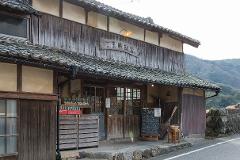 An old, traditional Japanese restaurant with a weathered wooden facade and a tile roof, displaying a menu board and vintage soda bottles, set against a backdrop of rolling hills.