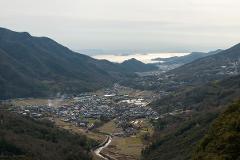 A scenic view of a valley with a town nestled between mountains, leading towards a body of water with islands in the distance under an overcast sky.