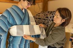 Experience the Excellence of Japanese Silk: Handwoven Ina Tsumugi A woman in a blue striped kimono is having an obi tied by another woman in a green jacket.