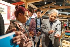 Experience the Excellence of Japanese Silk: Handwoven Ina Tsumugi An older Japanese man shows white silk threads to a group of people at an exhibition about Japanese silk.
