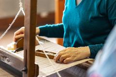 Experience the Excellence of Japanese Silk: Handwoven Ina Tsumugi A close-up of hands on a wooden loom, weaving fabric, with a person in a teal sweater in the background.