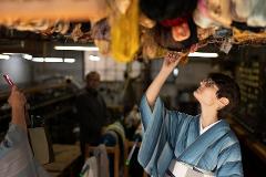 Experience the Excellence of Japanese Silk: Handwoven Ina Tsumugi A woman in a blue striped kimono examines silk threads hanging from the ceiling in a workshop.
