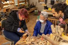 Experience the Craftsmanship of the Oroku Comb born in Kiso A woman works with a saw on a wooden block while an older man and a younger man watch in a woodworking shop.